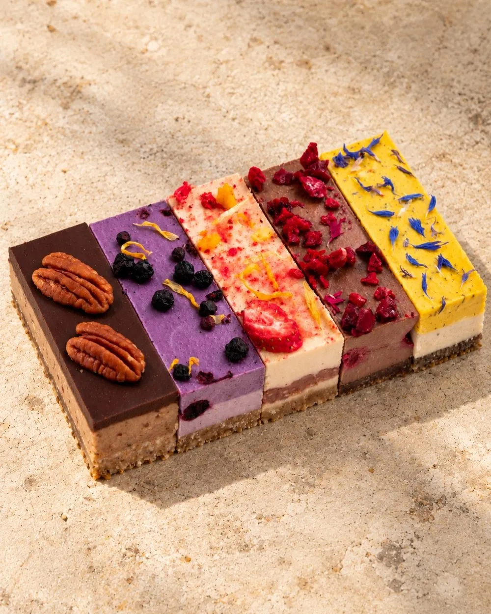 Five colourful raw cake slices side by side: pecan chocolate, blueberry, strawberry, raspberry and a yellow one topped with cornflower petals.